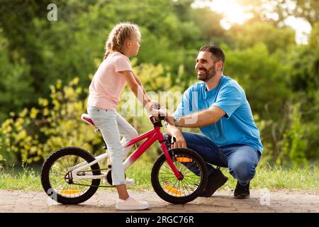 Glückliche Familienwerte. Seitlicher Blick auf den gutaussehenden Vater mittleren Alters und seine Tochter, die mit dem Fahrrad im grünen Park spazieren und reden Stockfoto