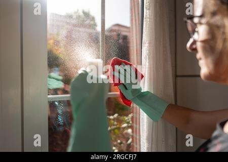 Frau, die Flüssigspray auf das Glas gießt, um es mit einem Tuch zu reinigen. Stockfoto