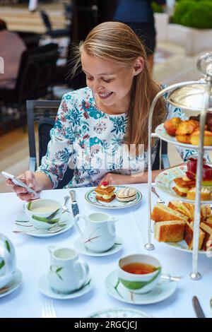 Wunderschöne junge Frau, die Nachmittagstee mit einer Auswahl an feinen Kuchen und Sandwiches in einem luxuriösen Pariser Restaurant genießt. Das Mädchen überprüft ihr Handy Stockfoto