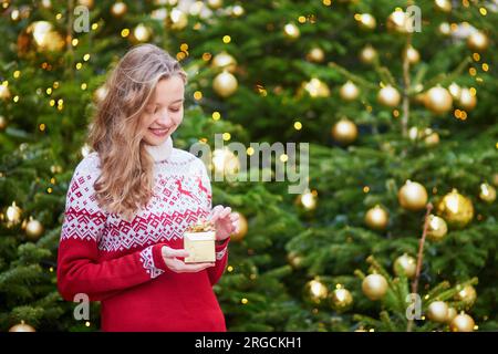 Fröhliches junges Mädchen mit einem kleinen Geschenkkasten in den Händen neben einem dekorierten Weihnachtsbaum, gelbe Weihnachtslichter im Hintergrund Stockfoto