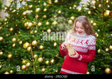 Fröhliches junges Mädchen öffnet kleine Geschenkschachtel in den Händen neben dem dekorierten Weihnachtsbaum, magische Glitzer erscheinen aus der Schachtel Stockfoto