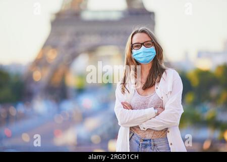 Ein Mädchen, das in der Nähe des Eiffelturms in Paris steht und eine Gesichtsschutzmaske trägt. Reiseurlaub in Frankreich während der Pandemie Stockfoto