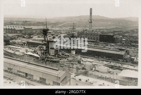 Rosyth Royal Naval Dockyard - Fife, Schottland - Panoramablick auf das Kraftwerk und den industriellen Schornstein mit mehreren Ziffernblättern. Im Vordergrund des Trockendocks befindet sich ein Schlachtschiff der Revenge-Klasse, wahrscheinlich HMS Resolution, das Flugzeugplattform auf dem Geschützturm zeigt. Stockfoto