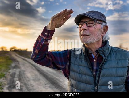 Ein älterer Bauer, der seinen Laptop in der Hand hält, auf einer unbefestigten Straße spaziert und jemanden in der Ferne begrüßt Stockfoto