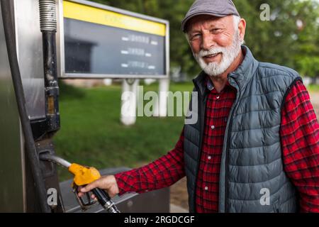 Senior-Mann in Karohemd mit Weste und Kappe, der die Treibstoffpistole auf einer kleinen Outdoor-Zapfsäule hält Stockfoto