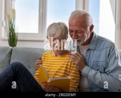 Fröhliches Seniorenpaar, das zu Hause auf dem Sofa sitzt, zusammen ein Buch liest und lächelt Stockfoto