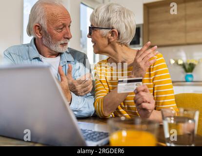 Fröhliches Seniorenpaar sitzt am Esstisch mit Küche im Hintergrund und kauft online mit Kreditkarte Stockfoto