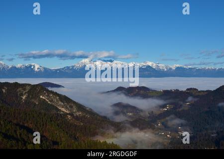 Blick auf die Berge Grintovec, Skuta und Kocna in den Kamnik-Savinja-alpen in Slowenien mit Nebel über den Bergen und zwischen den bewaldeten Hügeln Stockfoto