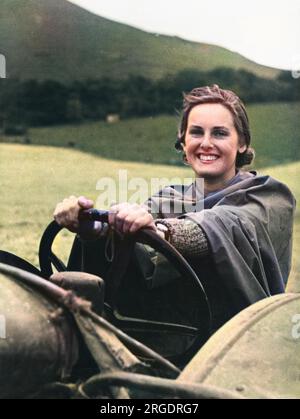 Land Girl, Lin Gapp, eine ehemalige Sekretärin in einem Kosmetiklabor, wird während des Zweiten Weltkriegs durch ein Bodenblech vor Regen geschützt, während sie einen Traktor auf einem Hof fährt Stockfoto