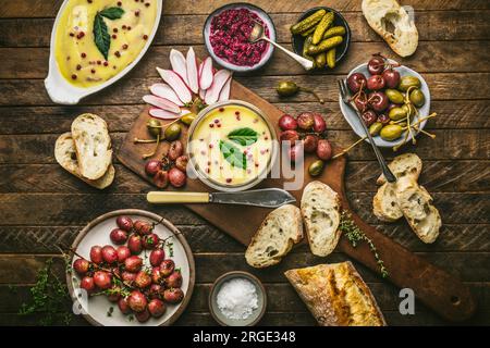 Becher mit Butter auf Holzbrett mit gerösteten Trauben, Radieschen, Gurken und Baguette Stockfoto