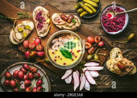 Pastete in Glasgefäß auf Holzbrett mit gerösteten Trauben, Radieschen, Gurken und Baguette Stockfoto