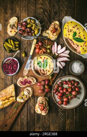 Pastete in Glasgefäß auf Holzbrett mit gerösteten Trauben, Radieschen, Gurken und Baguette Stockfoto