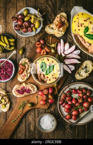 Pastete in Glasgefäß auf Holzbrett mit gerösteten Trauben, Radieschen, Gurken und Baguette Stockfoto