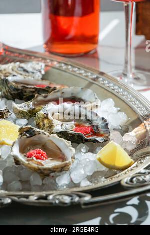 Frische Austern mit rotem Kaviar auf Eis in einer Silberschüssel und Rosenchampagner auf einem weißen Marmortisch Stockfoto