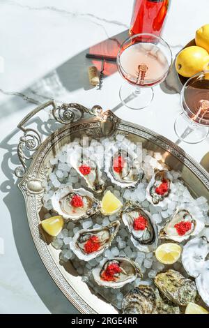 Frische Austern mit rotem Kaviar auf Eis in einer Silberschüssel und Rosenchampagner auf einem weißen Marmortisch Stockfoto