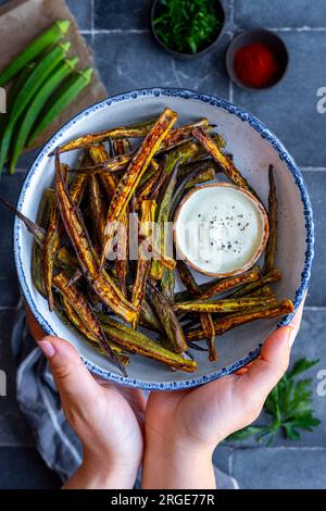 Frauenhände halten eine weiße Schüssel gefüllt mit gebratenem Okra und einer Dippsoße daneben. Stockfoto