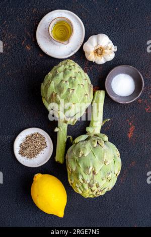 Artischocken, Olivenöl, Knoblauch, Salz, Pfeffer und Zitrone auf dunklem Hintergrund, fotografiert von oben. Stockfoto