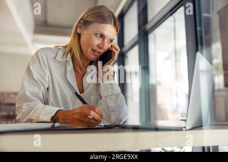 Unternehmerin, die sich Notizen macht und telefonisch mit dem Kunden spricht, während sie zusammenarbeitet Stockfoto