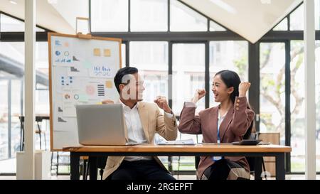Ein fröhlicher asiatischer Geschäftsmann zeigt seine Fäuste und feiert ein erfolgreiches Projekt mit einer Kollegin an einem Tisch im Büro. Stockfoto