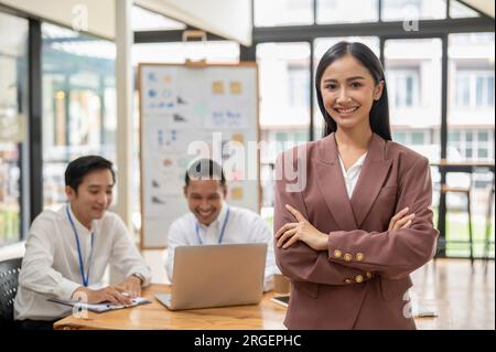 Eine selbstbewusste und umwerfende asiatische Geschäftsfrau oder CEO-Frau steht in einem Besprechungsraum mit verschränkten Armen, während ihre Mitarbeiter in einer Besprechung arbeiten Stockfoto