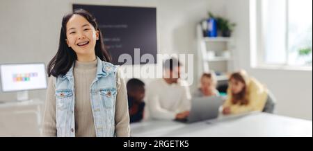 Der junge asiatische Schüler steht selbstbewusst in einem Programmierraum und schaut mit einem Lächeln in die Kamera, während der Lehrer andere Schüler im Hintergrund anspricht Stockfoto