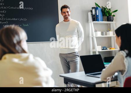 Junger männlicher Lehrer, der einen Programmierkurs für Kinder leitet. Computerwissenschaftslehrer steht vor der Klasse mit einem Lächeln, lehrend und führend Stockfoto