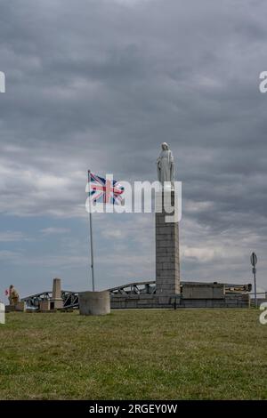 Arromanches-les-Bains, Frankreich - 07 21 2023 Uhr: Blick auf die Statue der Jungfrau Maria, englische Flagge und Denkmal für den D-Tag mit wolkigem Himmel über dem Stockfoto