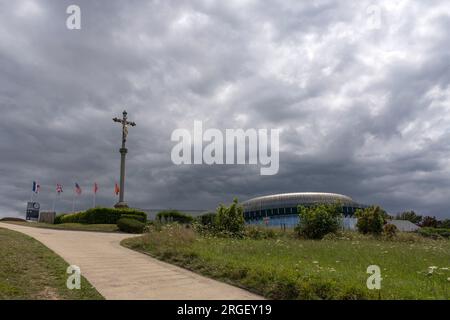 Arromanches-les-Bains, Frankreich - 07 21 2023: Blick auf das Kreuz Christi, Nationalflaggen und das kreisförmige Kino 360 mit wolkenlosem Himmel über der Kalvarien Stockfoto