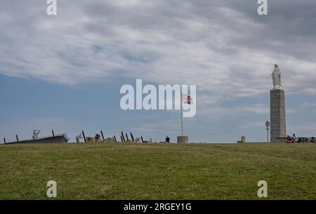 Arromanches-les-Bains, Frankreich - 07 21 2023 Uhr: Blick auf die Statue der Jungfrau Maria, englische Flagge und Denkmal für den D-Tag mit wolkigem Himmel über dem Stockfoto