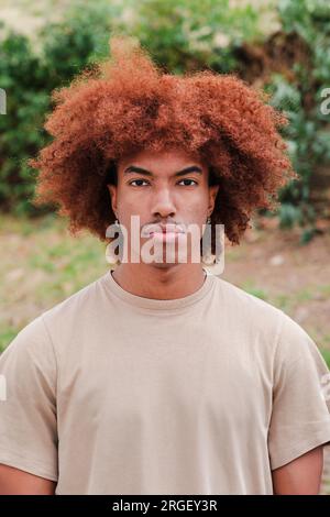 Vertikales Einzelbild eines jungen afroamerikanischen Mannes, der ernsthaft vor die Kamera schaut. Männlicher Teenager mit nachdenklicher Einstellung, der starrt. Vorderansicht eines rücksichtsvollen Typen mit Afrohaaren, der draußen steht. Hochwertiges Foto Stockfoto