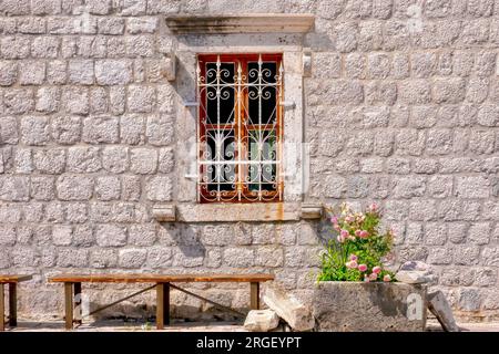 Ein malerisches Fenster mit kunstvoll verziertem schmiedeeisernem Gitter in einer alten Steinwand, mit einer Holzbank und pinkfarbenen Rosen davor. In Montenegro. Stockfoto