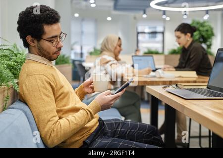 Seitliches Porträt eines jungen Mannes aus dem Nahen Osten, der eine Brille trägt, während der Pause im Büro Kaffee trinkt und Nachrichten abliest, Kopierraum Stockfoto