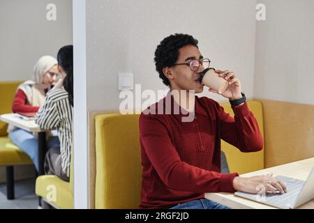 Porträt eines jungen Arabers, der Kaffee trinkt, während er einen Laptop an einer Kabine in einem Gemeinschaftsarbeitsplatz benutzt Stockfoto