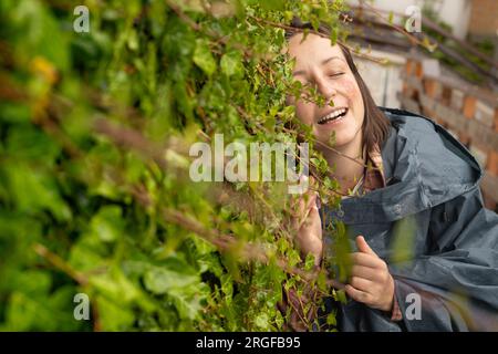 Junge Frau im Regenmantel steht gegen eine Efeu-Wand und genießt das Leben Stockfoto