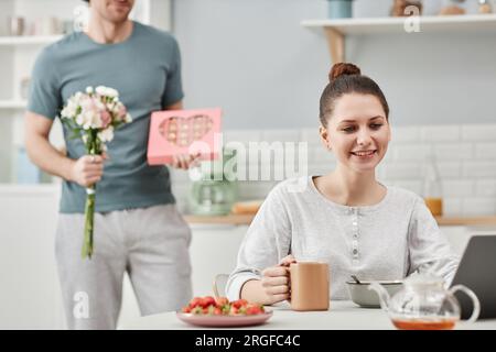 Porträt eines jungen Mannes, der seine Frau am Valentinstag mit Geschenken und Blumen überraschte, konzentriert sich auf eine lächelnde junge Frau, die im Vordergrund arbeitet Stockfoto
