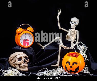 Halloween-Zeit. Ein Spielzeug-Skelett, das aus dem Grab kriecht und mit der Hand winkt, grüßt, Kürbisse mit Grimassen, eine orangefarbene Uhr, ein Totenkopf auf einem schwarzen Bac Stockfoto