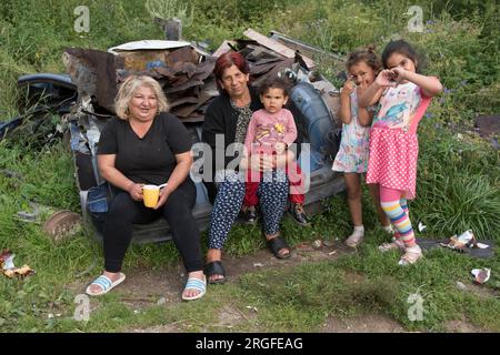 Romany Zigeunergroßmütter mit Enkelkindern. Sie sitzen auf einem verschrotteten Auto gegenüber von ihren Häusern. Die beiden Mädchen machen mit ihren Händen ein Herzzeichen und sehen ein Zeichen - Freundschaft. ZIGEUNER-Siedlung am Stadtrand von Sumiac, Bezirk Brezno, Slowakei, August 2023 2020s HOMER SYKES Stockfoto