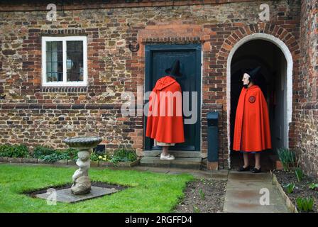 Die alten Damen von Castle Rising. Die Almeshouses - das Krankenhaus der heiligen und ungeteilten Dreifaltigkeit in Burgaufgang. Gegründet von Henry Howard, Earl of Northampton, gründete er eine Wohltätigkeitsorganisation, die 13 arme Jungfern oder Witwen aus der Gemeinde Castle Rising, die mindestens 50 Jahre alt war, beherbergte, von denen eine ihre Erzieherin war. Am Gründertag tragen sie ihre traditionellen roten Umhänge und spitzen schwarzen Hüte, um an einer Gedenkfeier in der „Krankenhaus“-Kapelle teilzunehmen. Die Dame öffnet ihre Haustür. Castle Rising, Norfolk, England, 24. Februar 2007. 2000s HOMER SYKES Stockfoto