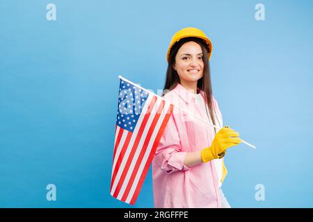 Ein Mädchen in einem Bauhelm und Handschuhen mit amerikanischer Flagge steht auf blauem Hintergrund Stockfoto