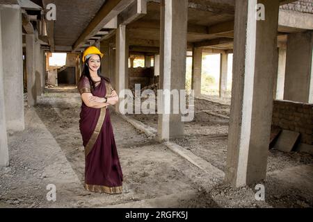 Porträt einer selbstbewussten jungen, wunderschönen indischen Bauingenieurin, die Saree und Helm trägt und auf der Baustelle Arme tragend steht. Weitwinkel, Kopieren Stockfoto