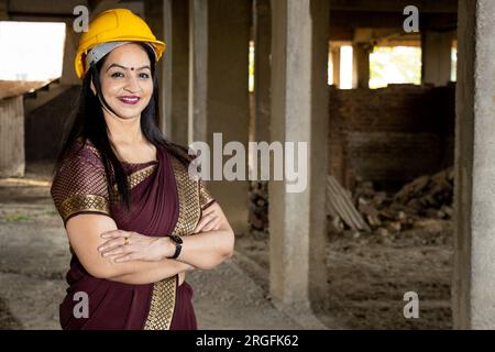 Porträt einer selbstbewussten jungen, wunderschönen indischen Bauingenieurin, die Saree und Helm trägt und auf der Baustelle Arme tragend steht. Kopierbereich, Stockfoto