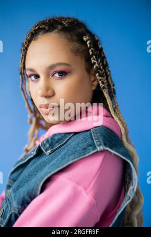 Attraktive afroamerikanische Frau mit Dreadlocks, die auf blauem Hintergrund auf die Kamera schaut, kühnes Make-up Stockfoto