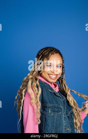 Porträt, fröhliche, dunkelhäutige Frau auf blauem Hintergrund, Jeansweste und rosafarbener Hoodie, Lächeln Stockfoto