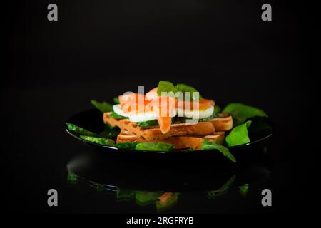 Gekochtes Sandwich mit leicht gesalzenem Rotfisch, Gurken und gekochten Eiern mit Kräutern, isoliert auf schwarzem Hintergrund. Stockfoto