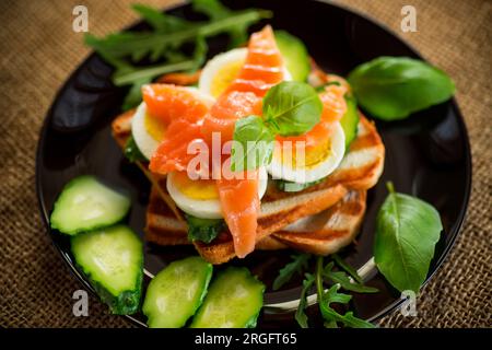 Gekochtes Sandwich mit leicht gesalzenem Rotfisch, Gurken und gekochten Eiern mit Kräutern, auf einem Teller. Stockfoto