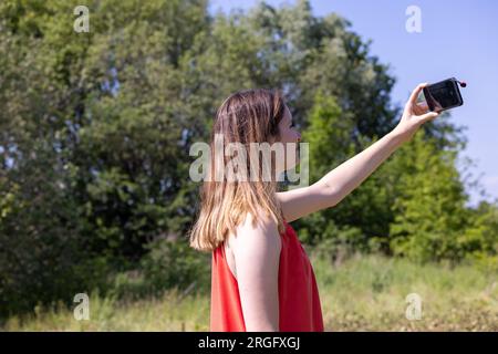 Eine malerische Szene, in der eine hübsche junge Kaukasierin in einem leuchtend roten Rock zu sehen ist, die einen perfekten Moment mit ihrem Mobiltelefon inmitten des üppigen grünen Waldes am Seeufer einfängt. Das strahlende Licht der Sonne verleiht der Umgebung einen Hauch von Magie, während sie die Schönheit der Natur verewigt und bleibende Erinnerungen schafft. Bilderperfekter Moment: Hübsche Frau, die Erinnerungen im Forest Lakeside einfängt. Hochwertige Fotos Stockfoto