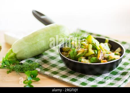 In Scheiben gebratene Zucchini mit Zwiebeln und Kräutern in einer Bratpfanne auf einem Holztisch. Stockfoto