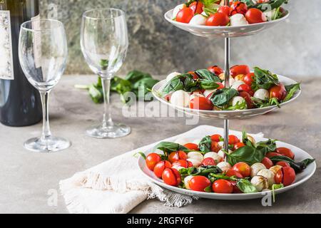 Klassischer italienischer Salat Insalata Caprese mit Kirschtomaten, Mini-Mozarella-Perlen, Basilikumblättern und Balsamglasur, serviert auf einem Kuchenstand, grauer Backg Stockfoto