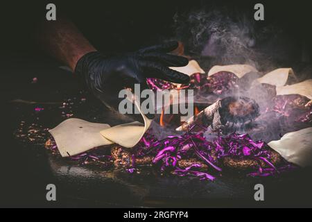 Ich bereite saftige Rinderhamburger mit Rotkohl zu. Grillfleisch brutzelt über heißen Flammen. Eine Hand streut Käse auf die Pasteten. Schwarzer Hintergrund. Stockfoto