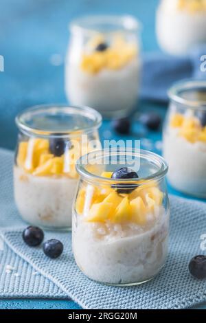 Kokosreis-Pudding mit Mango in Dessertgläsern auf blauem Hintergrund, vertikal Stockfoto